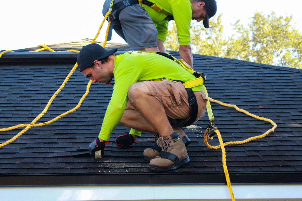 Two roofers in harnesses working on a dark shingle roof
