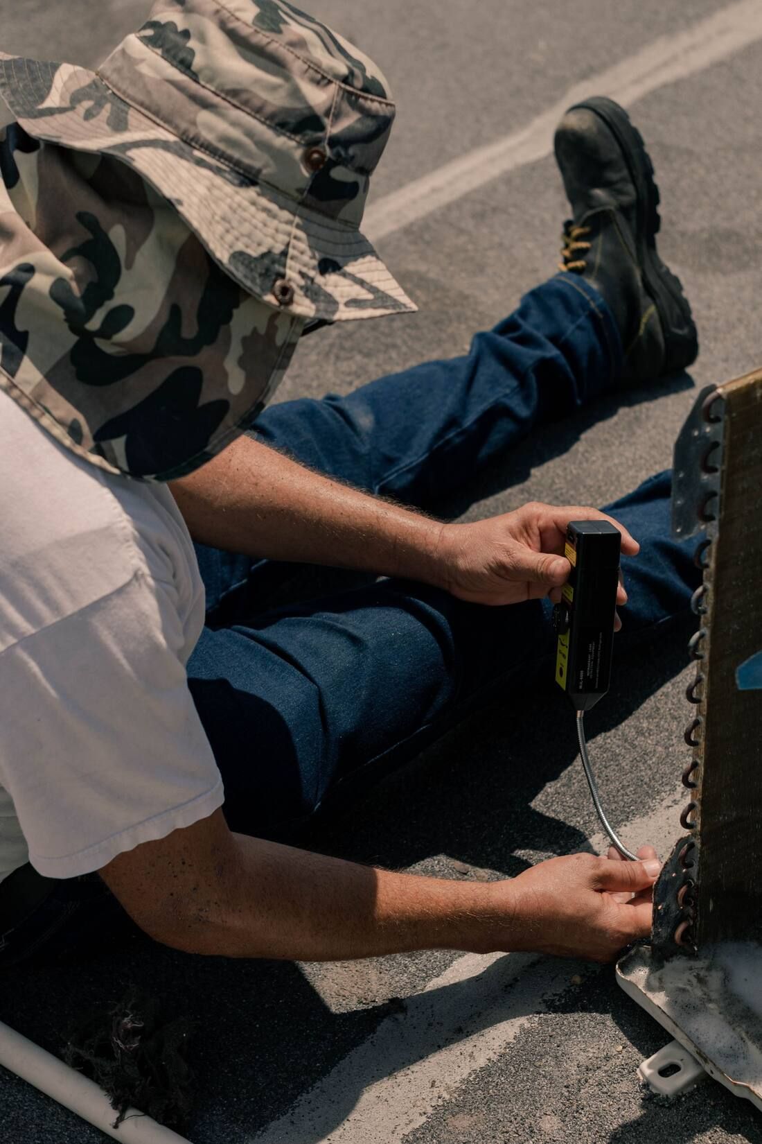 A technician in a bucket hat sitting beside an opened outdoor unit, checking its coil with a leak detector